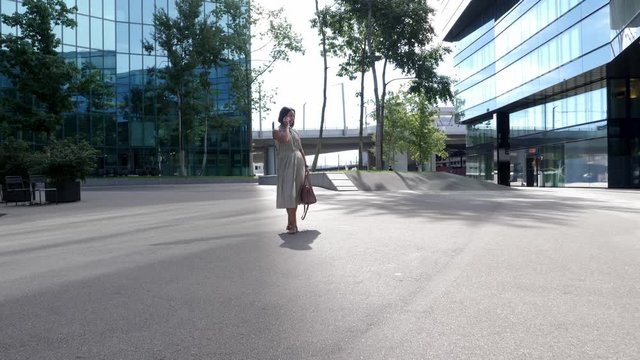 Young attractive businesswoman talks on phone in the bright sun between tall office buildings with glass facades. Slow Motion Wide Shot