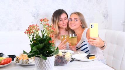 Obraz na płótnie Canvas friendship and party concept - two young beautiful girlfriends drink wine and take selfie on smartphone in a cozy kitchen at the table, selective focus