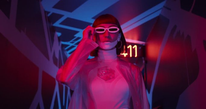 Attractive woman taking off sunglasses while walking in corridor with neon light and glowing numbers. Female model in futuristic outfit and make up looking to camera