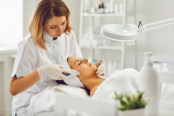 Young cosmetologist or dermatologist making ultrasound facial cleaning for woman in beauty salon