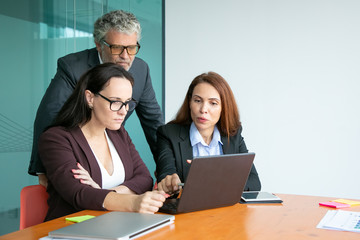 Business team watching presentation on laptop, pointing at display, discussing details while sitting and standing at meeting able together. Medium shot. Communication and teamwork concept