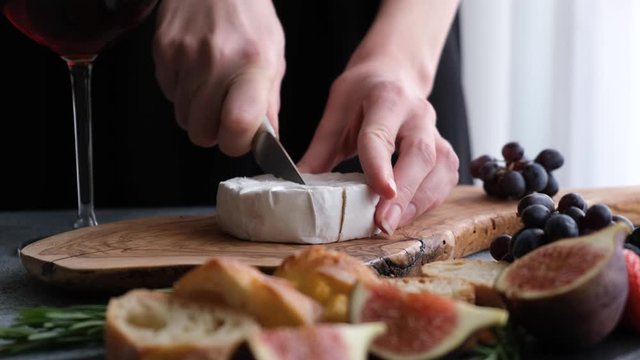 Cutting camembert cheese on wooden board. Preparation of cheese plate with fresh figs, grapes, baguette cheese and wine