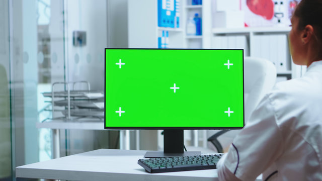 Female doctor working on computer with green screen in private clinic cabinet. Assistant in uniform. Medic in white coat working on monitor with chroma key in clinic cabinet to check patient diagnose.