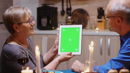 Old retired senior couple using green mock-up screen digital isolated tablet computer. Aged people looking at green screen template chroma key display sitting at the table in kitchen during dinner.