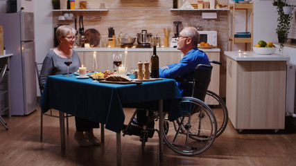 Senior couple coming home for romantic dinner. Old man in wheelchair dining with his cheerful wife sitting at the table in the kitchen. Imobilized paralyzed handicapped husband having romantic dinner