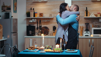 Young caucasian man cooking for his wife a romantic dinner, waiting in kitchen. Preparing festive dinner with healty food, woman coming back from job surprised kissing and hugging her husband.