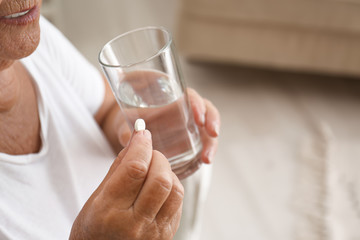 Senior woman with glass of water taking pill at home, closeup