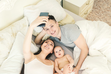 Happy young father, mother and cute baby girl lying on light bed and make selfie. Top view.