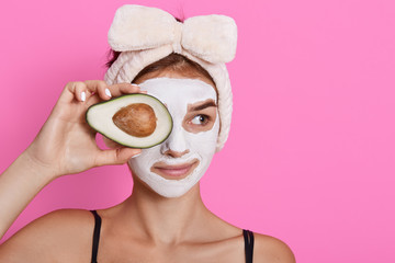 Young woman holding avocado in hands and covering her eyes with fruit, having white mask on face, looking aside, wearing head band with bow isolated over rose background.