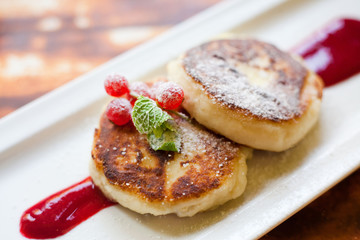 Cheesecakes sprinkled with powdered sugar decorated with currants and jam on a rectangular plate, served with sour cream