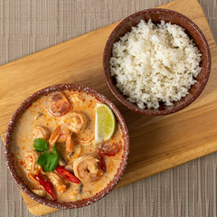 Ceramic bowl of Thomas soup with shrimp and coconut milk and a bowl of rice on the background of wooden boards and a gray mat. Asian food. Top view.