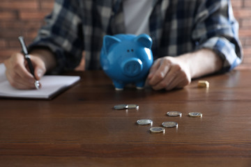Man with piggy bank counting money at wooden table, focus on scattered coins