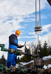 Engineer in work overalls and orange helmet checking oil pumping unit at oil field, using laptop. Male worker standing in front of oil well pump jack. Concept of petroleum industry, oil extraction.
