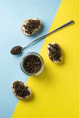 Glass jar and spoon of black caviar and sandwiches with black caviar on blue and yellow background. Top view and copy space