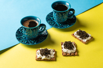 Coffee in blue cups and crackers with cream cheese and black caviar on a blue and yellow background.