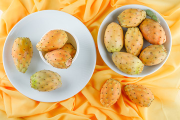 Cactus fruits in bowl and plates on yellow textile background, flat lay.