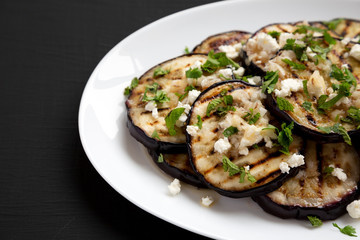 Homemade Grilled Eggplant with Feta and Herbs on a white plate on a black surface, side view. Copy space.