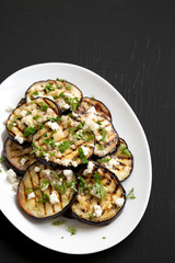 Homemade Grilled Eggplant with Feta and Herbs on a white plate on a black background, high angle view. Copy space.