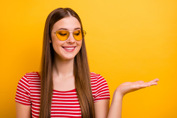 Photo of lovely pretty young girl smiling hand hold look empty space choosing better variant advising object wear sun specs striped white red shirt vibrant yellow color background