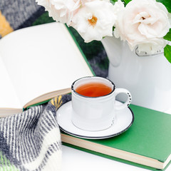 Cup of tea near open book, flowers white wild rose in vase, warm plaid on white table outside in garden. Romantic leisure breakfast with nature background. Cozy home backyard, reading in provence cafe