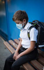 A child of the European race sits sad on a bench with a knapsack and a protective medical mask. A schoolboy goes to school in the midst of the COVID-19 pandemic. Go to school wearing masks.