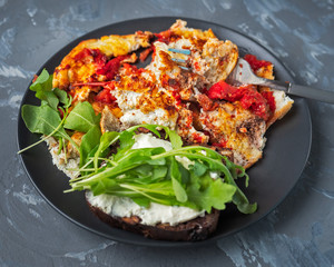 Fried eggs with tomatoes and fresh arugula for a quick breakfast on a dark gray plate, top view on a gray background