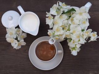 Herbal aromatic tea in a white сup with a saucer, mug with milk and a branch of Jasmine with white flowers in kteakettle on a wooden background , top view.   Flower still life with a traditional drink