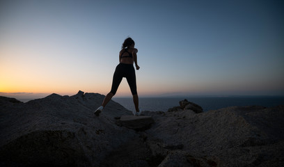 An young active sporty athlete woman with firm fit body is making running and jogging workout on a top of rock with seascape at sunset.