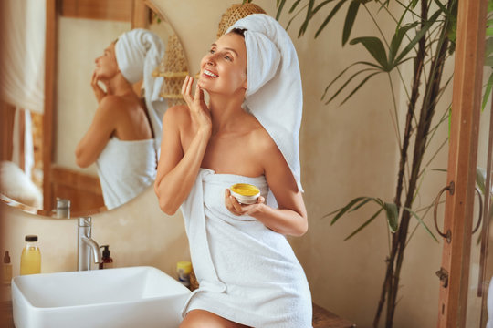 Woman After Bath. Portrait Of Happy Model Wearing Towel After Shower. Young Female Applying Anti-Aging Cream Or Mask On Facial Skin. Natural Beauty Product For SPA Treatment At Home.