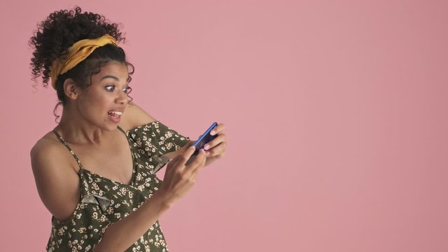 A happy african american young woman wins the game on her smartphone standing near free space isolated over pink background