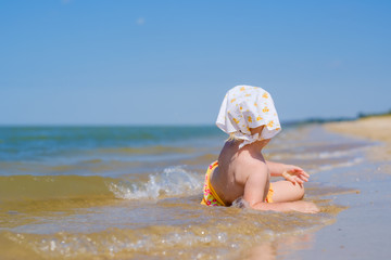 Selective focus on baby girl lying in the water on the sand on the beach. Sunny day. Back view. On the head is a white bandana shawl. Shallow depth of field. Happy childhood. Copy space.