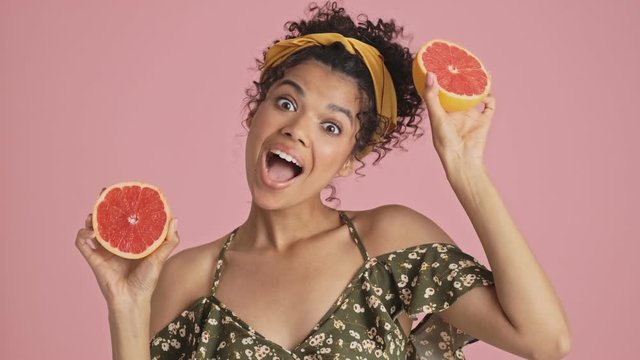 A close-up view of a cheerful african american young woman is posing with grapefruits standing isolated over pink background