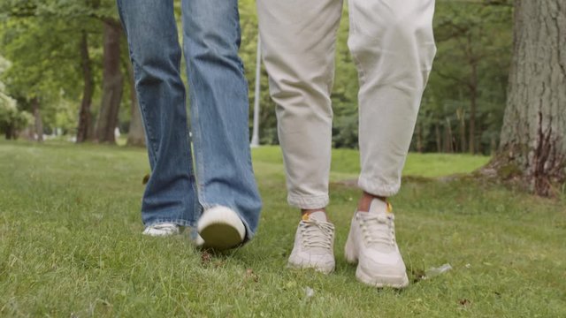 Close up of legs of unrecognizable man and woman wearing jeans and sneakers are walking on grass in park