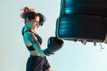 Athletic female fighter trains uppercut on punching bag made of tires in studio in neon light....