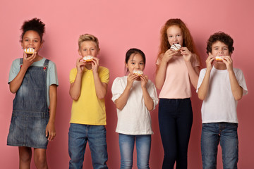 Cute diverse children eating yummy donuts over pink background