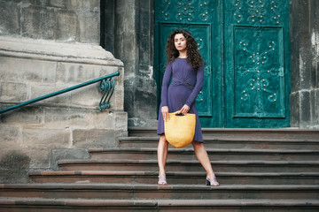 Fototapeta na wymiar Young beautiful brunette woman wearing purple polka dot dress, holding knitted yellow bag standing on the stairs posing in street of city. Copy, empty space for text