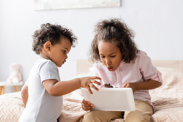 Fototapeta premium surprised african american girl using digital tablet near little brother in bedroom