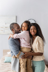 african american man holding daughter on hands while mother hugging them and looking at camera