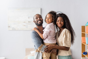 bearded african american man holding daughter near mother hugging them and looking at camera