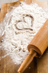 Love baking concept - rolling pin with heart shape in flour on wooden cutting board.