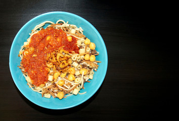 Famous traditional Arabic, Egyptian dish - Koshari. National Kushari in a turquoise plate on dark wooden background with copy space. Flat lay, top view.