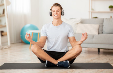 Fototapeta na wymiar Modern workout with music at home. Smiling guy in headphones enjoys yoga and meditation