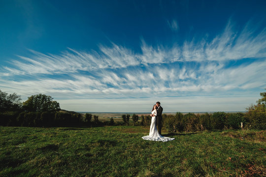 elegant groom hugs a gorgeous brunette bride on a background of nature and blue sky