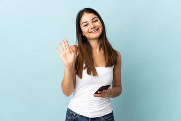 Young caucasian woman using mobile phone isolated on blue background saluting with hand with happy expression
