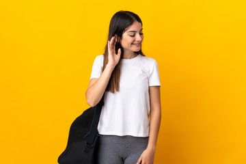Young sport woman with sport bag isolated on yellow background listening to something by putting hand on the ear