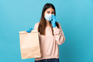 Young woman protecting from the coronavirus with a mask and holding a grocery shopping bag isolated on blue background shouting with mouth wide open