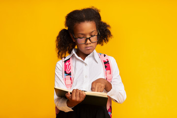 African Schoolgirl Learning To Read Holding Book Standing In Studio - obrazy, fototapety, plakaty