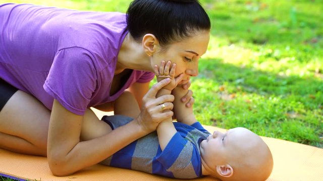 Jovial mother playing with cute baby lying on soft mat in park. Young woman in sportswear kissing and clapping hands of child. Concept of maternity