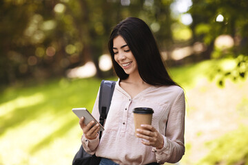 Fototapeta premium Asian Student Girl Using Smartphone While Walking With Backpack And Coffee Outdoors
