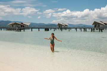 Fototapeta na wymiar Young slim tourist woman enjoy suntan, standing in the tourquise water in front of wooden owrwater bungalows, wearing swimsuit. Vacation concept